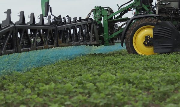 An eye-level shot of a John Deere See & Spray™ system in a field. A black boom arm extends toward a large yellow tractor wheel, releasing targeted bursts of vibrant blue mist onto green crops. The image demonstrates precision agriculture by spraying only specific areas rather than the entire field.