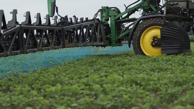 An eye-level shot of a John Deere See & Spray™ system in a field. A black boom arm extends toward a large yellow tractor wheel, releasing targeted bursts of vibrant blue mist onto green crops. The image demonstrates precision agriculture by spraying only specific areas rather than the entire field.