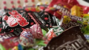 A close-up, slightly high-angle photograph shows a colorful, haphazard pile of various classic American candies. The foreground is dominated by a black and red pouch of Strawberry Pop Rocks popping candy. To its right, a green and yellow six-piece pack of Now and Later chewy candy is visible. Scattered throughout the mix are several Charms Blow Pops in bright red and blue wrappers, along with a pink-wrapped Dum Dums lollipop labeled "Bubble Gum." Other partially obscured candy wrappers in shades of red, yellow, and green create a vibrant, textured background. The lighting is soft, and the shallow depth of field keeps the central candies in sharp focus while the edges of the pile are softly blurred.