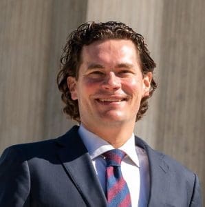 A head-and-shoulders portrait of a man with light skin, dark brown curly hair, and a warm smile. He is wearing a dark navy suit jacket over a crisp white dress shirt and a red and navy diagonally striped tie. The lighting is bright and natural, casting soft shadows on his face, against a blurred, light gray architectural background.