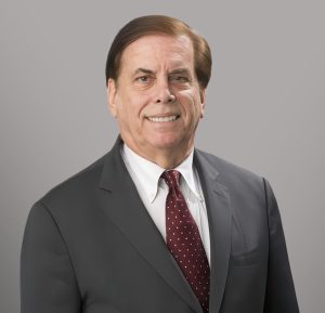 A professional headshot of a middle-aged man with fair skin and dark brown hair that is parted to the side. He is smiling, showing his teeth, and looking directly at the camera. He is wearing a dark grey suit jacket over a crisp white dress shirt and a maroon necktie with small white polka dots. The background is a solid, neutral light grey.