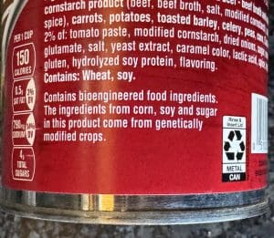 A close-up of a red Campbell’s soup label. White text lists ingredients like beef broth, barley, and vegetables. A "Contains Bioengineered Food Ingredients" disclosure is prominent. A sidebar shows 150 calories and 790mg sodium per serving. A "How2Recycle" metal can icon is in the corner.