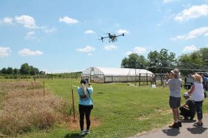 An agricultural spray drone hovers in a blue sky over an open field during a farming demonstration. A woman in a blue shirt uses her phone to take a photo of the drone from behind. On the right, a small group of people watch and film the flight. In the background, a large white hoop house greenhouse stands on the green grass near a wire fence and distant trees.