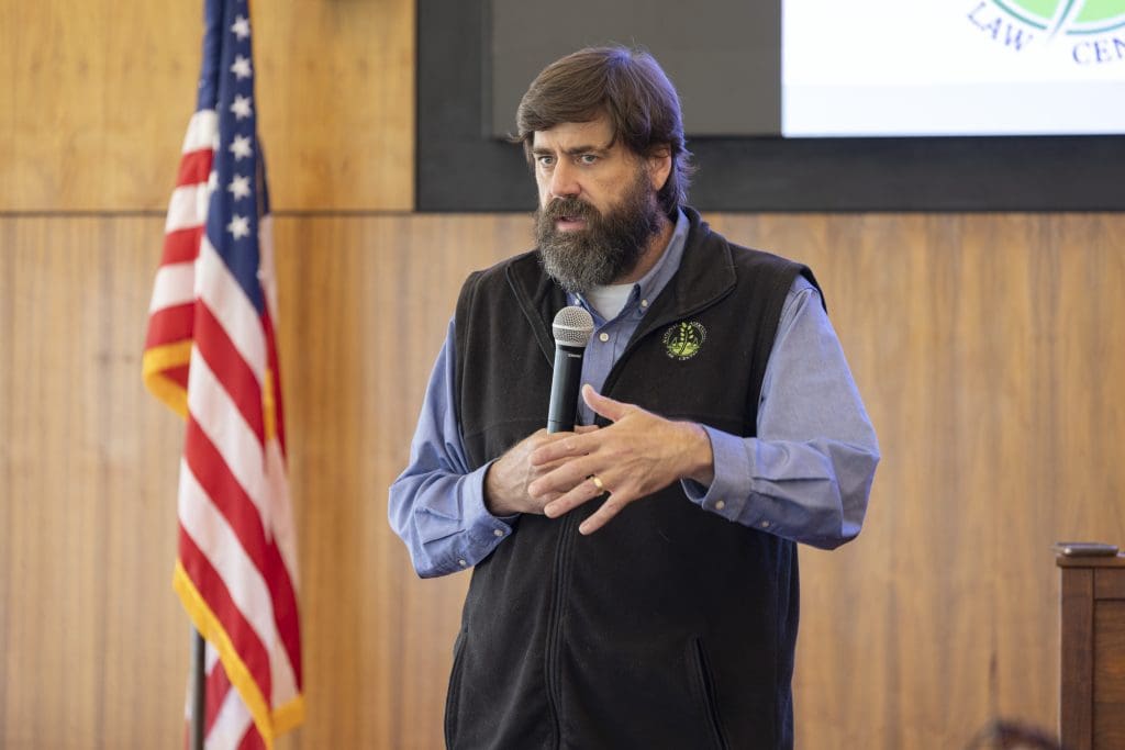 A man with a full, dark beard and brown hair speaks into a microphone held in his right hand. He wears a dark sleeveless zip-up vest over a light-blue button-down shirt. His left hand is raised to his chest. Behind him, an American flag stands on the left, and a wooden paneled wall serves as the background, with part of a screen visible on the top right showing a green logo with the words "LAW CENTER."
