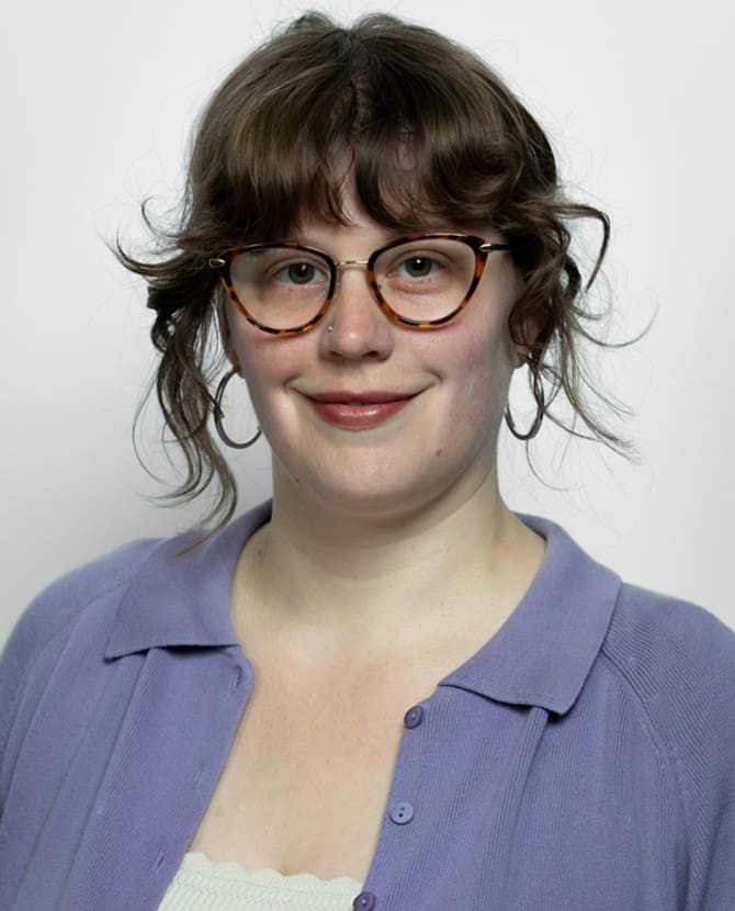 The image displays a woman with a distinct personal style featuring a textured brunette shag haircut with wispy bangs. She is wearing tortoiseshell cat-eye glasses frames, a small nostril piercing, silver hoop earrings, and a lavender-purple knit cardigan with a polo collar.