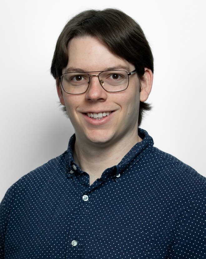 A professional headshot of a young adult with light skin, short brown hair styled with a middle part, and thin-rimmed metal glasses. They are smiling broadly, showing their teeth, and looking directly at the camera. They are wearing a dark blue, long-sleeved button-down shirt patterned with small white polka dots. The background is a solid, clean off-white. The lighting is bright and even, highlighting their face clearly.