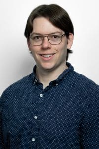 A studio portrait of a young adult with light skin and short, straight dark-brown hair parted down the middle.The individual is smiling, showing their teeth, and looking directly at the camera. They are wearing thin-rimmed, gold aviator-style glasses and a navy-blue, long-sleeved button-down shirt featuring a white polka-dot pattern. The background is a solid, clean white. The lighting is bright and even, cast from the front left.