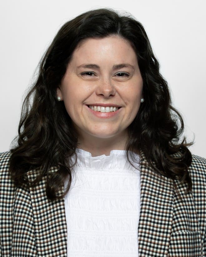 A professional studio headshot of a smiling woman with medium-length, dark, wavy hair parted in the middle. She is wearing small pearl earrings, a white high-neck blouse with a ruffled collar, and a brown, black, and white houndstooth plaid blazer. The background is a solid, neutral light gray.